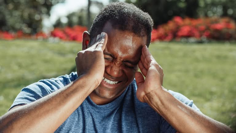 Adult man outdoors with hands on head showing signs of discomfort or headache.